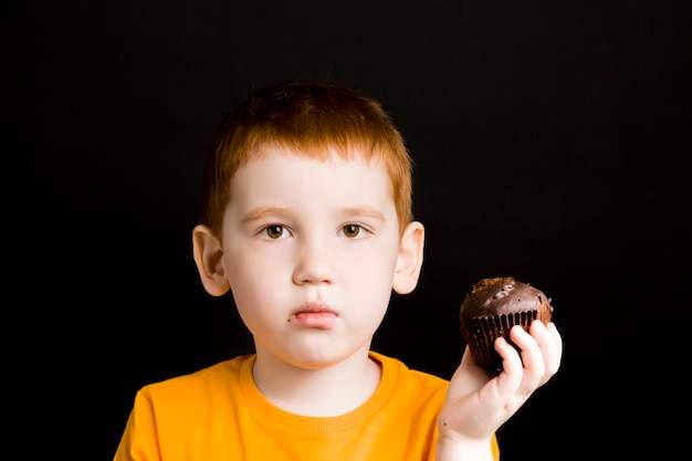 rothaariger Junge mit einem Schokoladencupcake, ein Junge isst einen Cupcake mit Vergnügen, süßes schädliches, aber leckeres Essen bei einem Kind