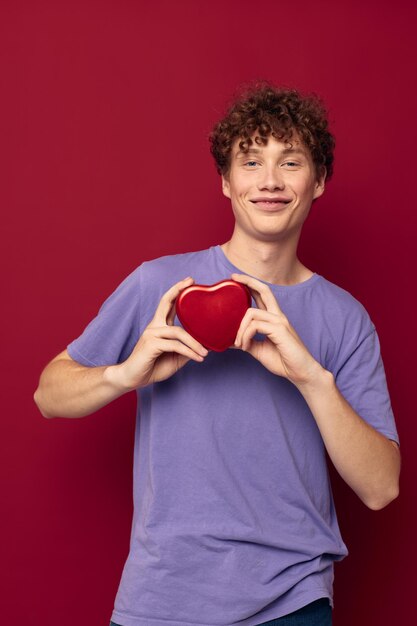Rothaariger lockiger Kerl eine Schachtel in Form eines Herzens in seinen Händen auf rotem Hintergrund