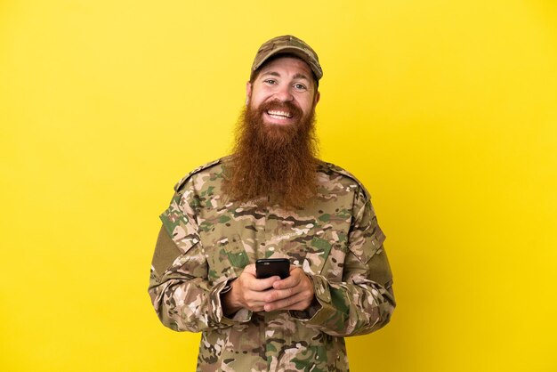 Rothaariger Mann des Militärs über isoliert auf gelbem Hintergrund, der eine Nachricht mit dem Handy sendet