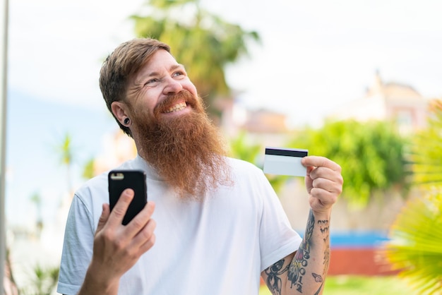 Rothaariger Mann mit Bart, der beim Denken mit dem Handy mit einer Kreditkarte kauft