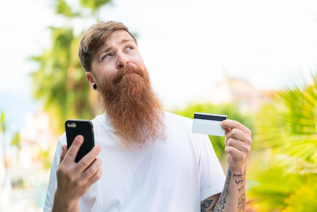 Foto rothaariger mann mit bart, der beim denken mit dem handy mit einer kreditkarte kauft