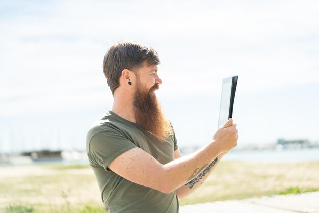 Rothaariger Mann mit Bart, der draußen ein Tablet mit glücklichem Gesichtsausdruck hält