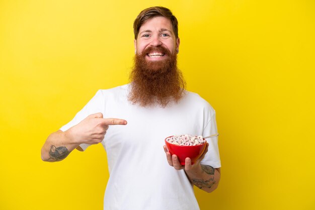 Rothaariger Mann mit Bart, der eine Schüssel Müsli isst, isoliert auf gelbem Hintergrund und darauf zeigt