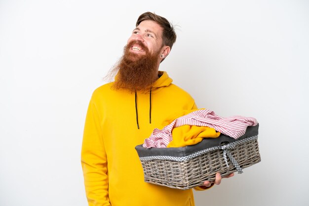 Rothaariger Mann mit Bart, der einen Kleiderkorb isoliert auf weißem Hintergrund hält und eine Idee denkt, während er nach oben schaut