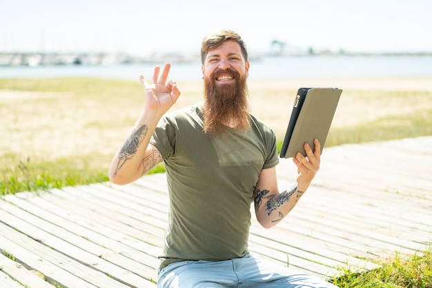 Foto rothaariger mann mit bart, der im freien ein tablet hält und mit den fingern ein ok-zeichen zeigt