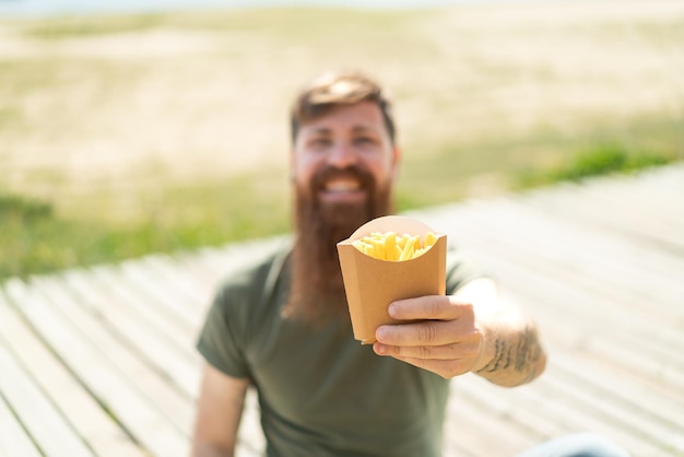 Rothaariger Mann mit Bart, der im Freien gebratene Chips mit fröhlichem Gesichtsausdruck hält