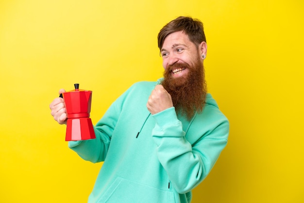 Rothaariger Mann mit Bart, der Kaffeekanne isoliert auf gelbem Hintergrund hält und einen Sieg feiert