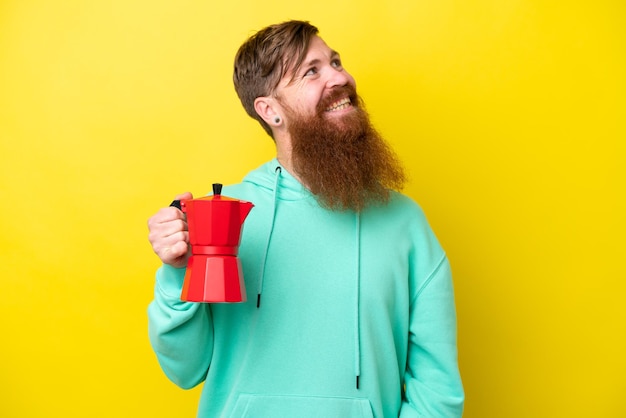 Rothaariger Mann mit Bart, der Kaffeekanne isoliert auf gelbem Hintergrund hält und lächelnd nach oben schaut