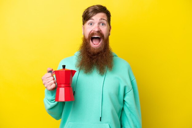 Rothaariger Mann mit Bart, der Kaffeekanne isoliert auf gelbem Hintergrund mit überraschtem und schockiertem Gesichtsausdruck hält