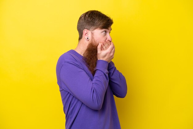 Rothaariger Mann mit Bart isoliert auf gelbem Hintergrund, der den Mund bedeckt und zur Seite schaut