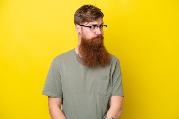 Rothaariger Mann mit Bart isoliert auf gelbem Hintergrund, der Zweifel hat, während er zur Seite schaut