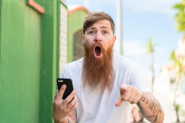 Rothaariger Mann mit Bart mit Handy im Freien überrascht und nach vorne zeigend