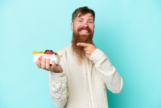 Rothaariger Mann mit langem Bart, der eine Obstschale isoliert auf blauem Hintergrund hält und darauf zeigt