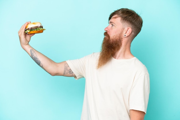 Rothaariger Mann mit langem Bart, der einen Burger isoliert auf blauem Hintergrund mit glücklichem Ausdruck hält
