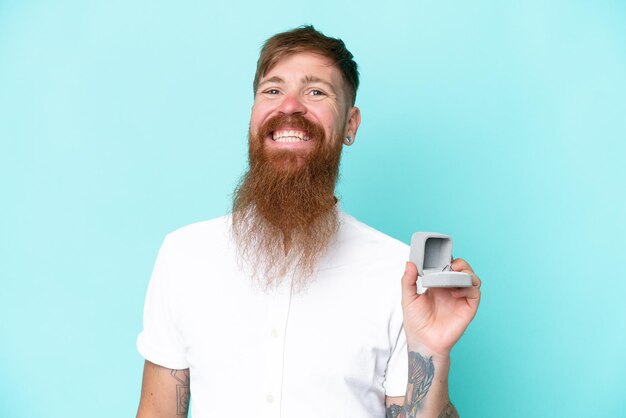 Rothaariger Mann mit langem Bart, der einen Verlobungsring isoliert auf blauem Hintergrund hält und viel lächelt