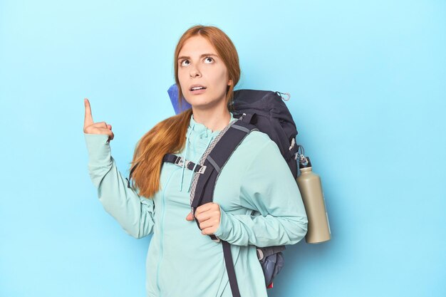 Rothaariger Reisender mit Rucksack und Matte im Studio, der mit geöffnetem Mund nach oben zeigt