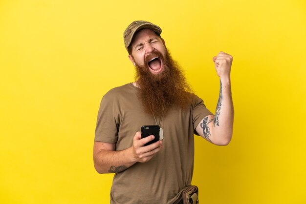 Rothaariger Soldat mit Erkennungsmarke auf gelbem Hintergrund mit Telefon in Siegesposition