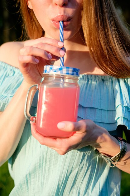 Rothaariges Ingwermädchen gekleidet im blauen Kleid trinkt rosa Smoothie vom Glasbecher im Sommer