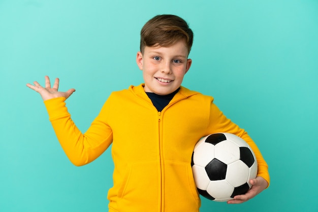 Rothaariges Kind, das Fußball auf blauem Hintergrund mit schockiertem Gesichtsausdruck spielt