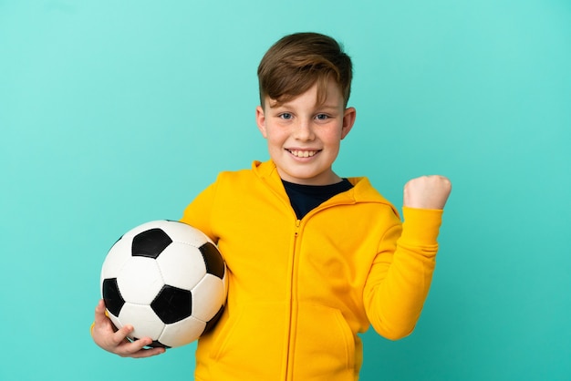 Rothaariges Kind spielt Fußball isoliert auf blauem Hintergrund und feiert einen Sieg in Siegerposition winner