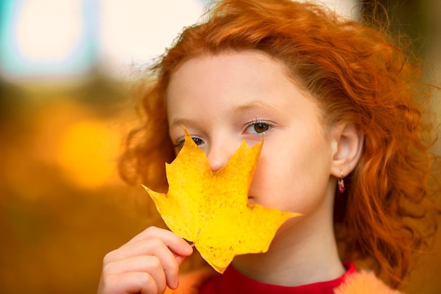 Rothaariges kleines Mädchen schließt ein Auge mit einem gelben Ahornblatt Trauriges Kind an einem Herbsttag in der Nähe