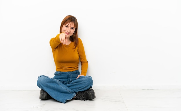 Rothaariges Mädchen, das auf dem Boden sitzt, isoliert auf weißem Hintergrund, zeigt mit einem selbstbewussten Ausdruck mit dem Finger auf dich