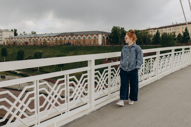 Foto rothaariges mädchen in jeanskleidung geht entlang der stadtbrücke