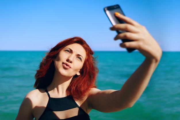 Rothaariges Mädchen lässt ihre Lippen sich ducken und macht Selfie auf der Smartphone-Kamera hübsche Frau in schwarzen Schwimmbädern...