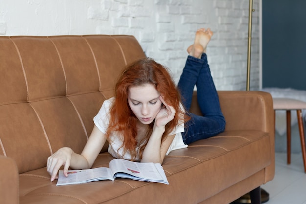 Rothaariges Mädchen liegt auf der Couch und liest zu Hause eine Zeitschrift