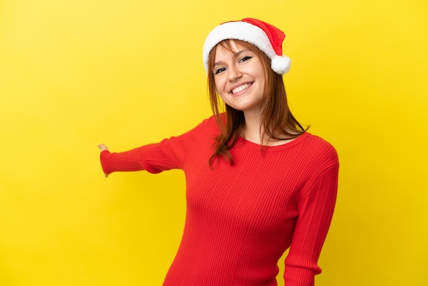 Rothaariges Mädchen mit Weihnachtsmütze isoliert auf gelbem Hintergrund, das die Hände zur Seite ausstreckt, um einzuladen, zu kommen