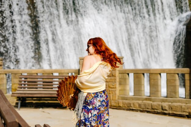 Rothaariges, stilvolles Mädchen geht in der Sonne auf dem Hintergrund eines Wasserfalls durch den Park
