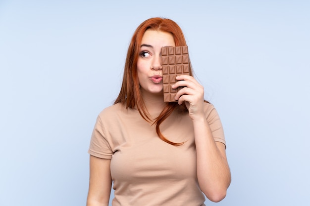 Rothaariges Teenager-Mädchen über Blau, das eine Schokoladentafel nimmt und überrascht