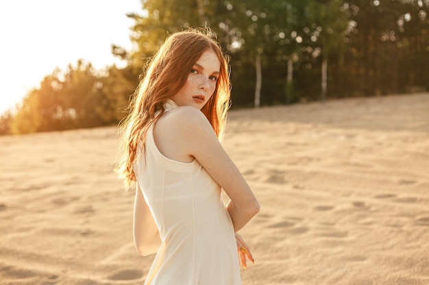 rothaariges weibliches Modell mit Sommersprossen und langen Haaren, die im Sommer am Sandstrand stehen