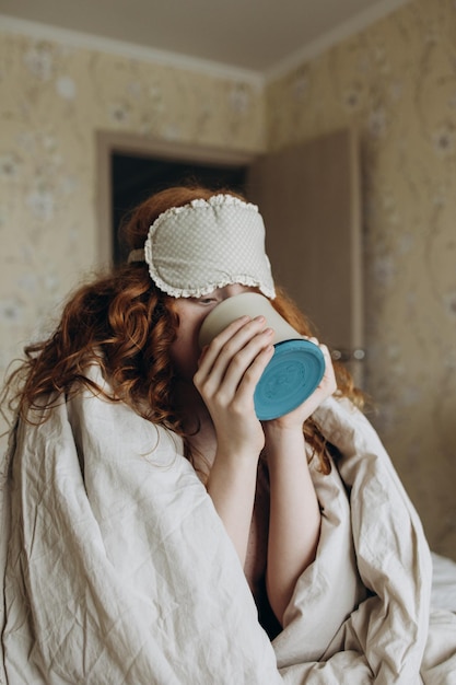 Rothaariges, zotteliges Mädchen wickelte sich in eine Decke und trinkt Kaffee, während es auf dem Bett sitzt
