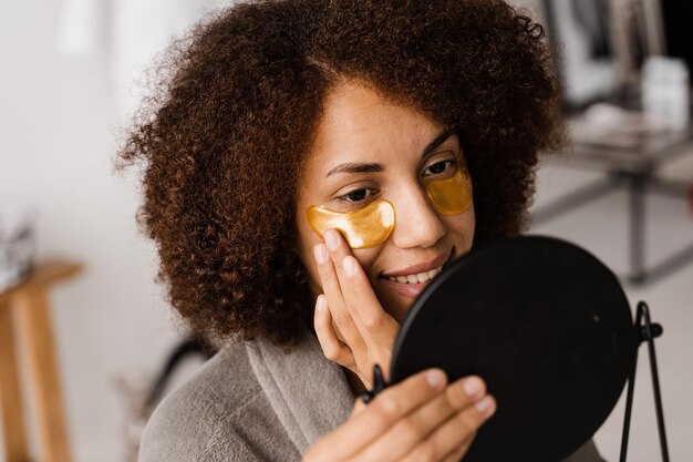 Rotina de beleza matinal Mulher afro-americana com manchas douradas nos olhos olha no espelho para sua pele Garota africana em roupão de banho aplica manchas douradas nos olhos no banheiro