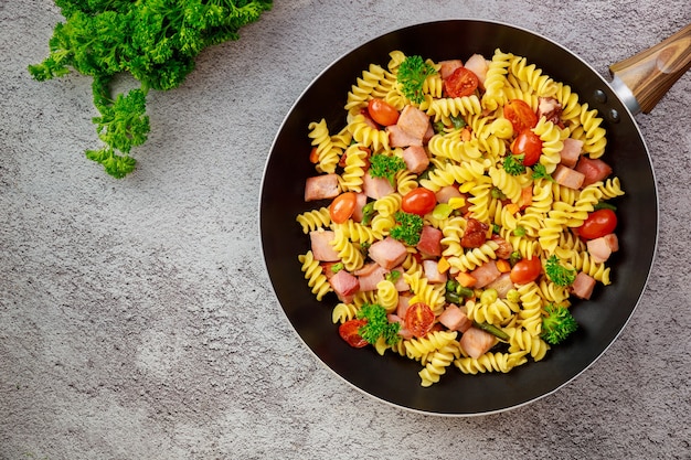 Rotini de macarrão colorido, tomate cereja e presunto na panela. Vista do topo.