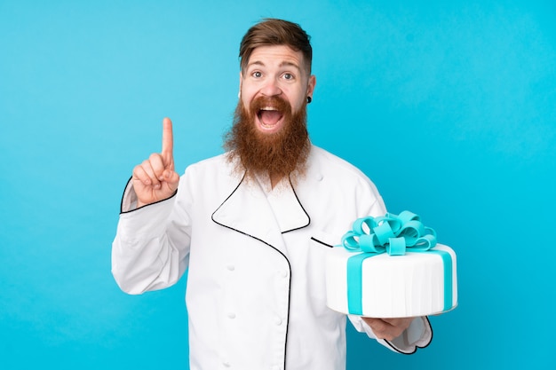 Rotschopf-Konditor mit langem Bart, der einen großen Kuchen über isolierter blauer Wand hält, die eine große Idee aufzeigt