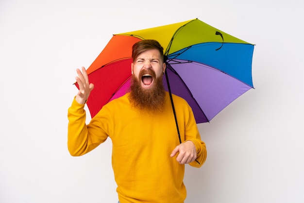 Rotschopf Mann mit langem Bart hält einen Regenschirm über isolierte weiße Wand unglücklich und frustriert mit etwas