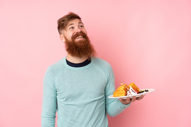 Rotschopfmann mit langem Bart, der Waffeln über isolierter rosa Wand hält, die beim Lächeln nach oben schaut