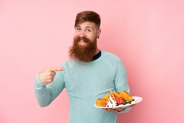 Foto rotschopfmann mit langem bart hält waffeln über isolierter rosa wand und zeigt darauf