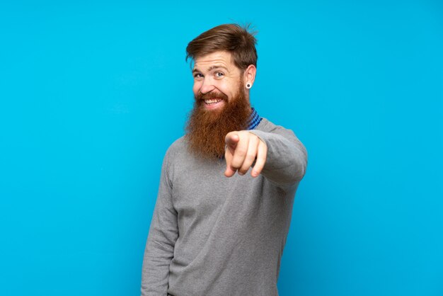 Rotschopfmann mit langem Bart über blauer Wand zeigt mit einem selbstbewussten Ausdruck auf Sie