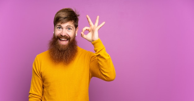 Rotschopfmann mit langem Bart über isoliertem Purpur überrascht und zeigt ok Zeichen