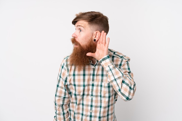 Rotschopfmann mit langem Bart über isolierter weißer Wand, der etwas hört, indem er Hand auf das Ohr legt