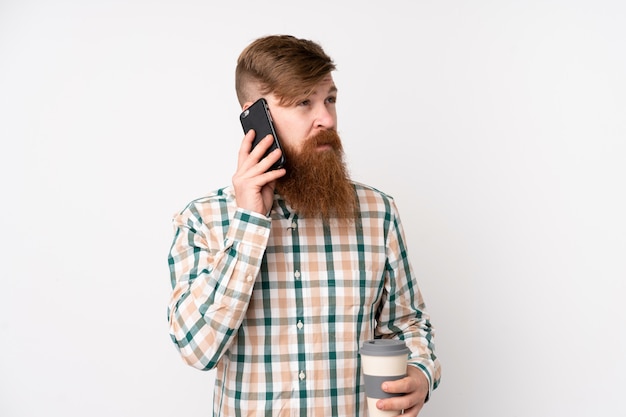 Rotschopfmann mit langem Bart über isolierter weißer Wand, die Kaffee zum Mitnehmen und ein Handy hält