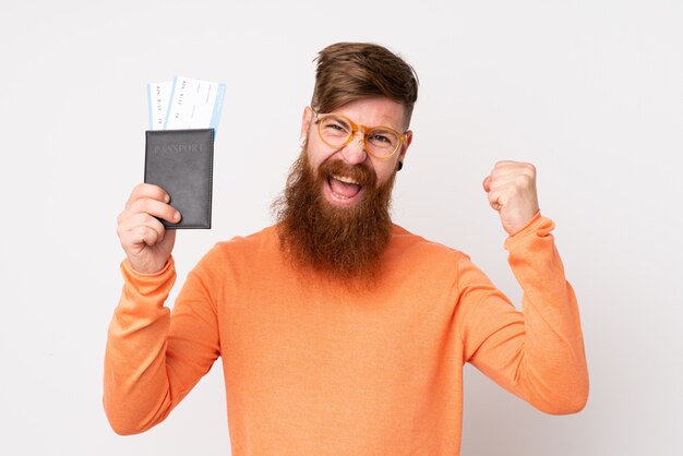 Rotschopfmann mit langem Bart über isolierter weißer Wand glücklich im Urlaub mit Pass und Flugtickets