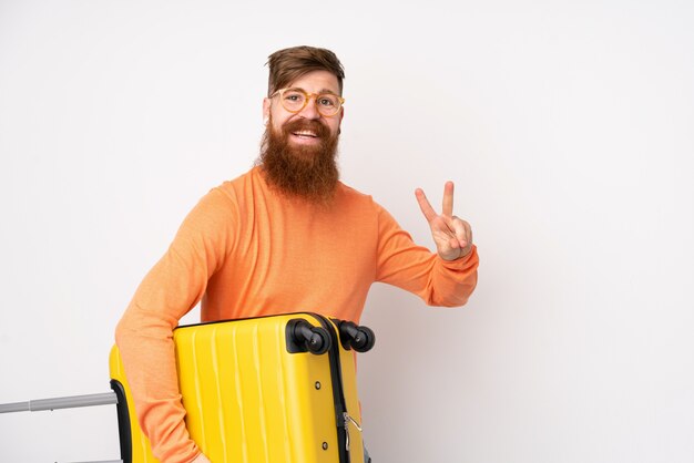 Rotschopfmann mit langem Bart über isolierter weißer Wand im Urlaub mit Reisekoffer und Siegesgeste