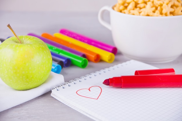 Rotuladores de colores, cuaderno con scetch, tazón de cereal en forma de estrella y manzana sobre el fondo gris