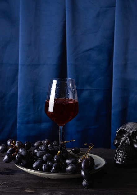 Rotwein in einem Glasglas, Trauben auf einem Teller und Teil eines Schädels auf blauem Stoffhintergrund. Das Konzept der Magie, Mystik und Esoterik.