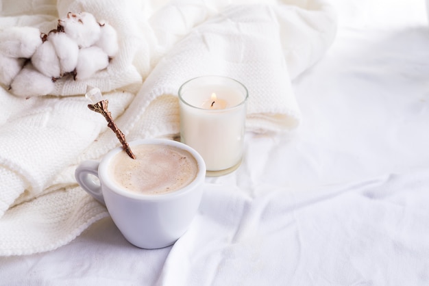 Roupa de cama com uma manta de malha fofa e xícara de café, flores de algodão e velas.