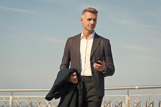 Roupa formal de empresário com smartphone Conceito de vida moderna Chefe a caminho do escritório Use telefone enquanto caminha Estilo urbano Dia de trabalho regular Homem carismático fundo de céu azul Chamada telefônica